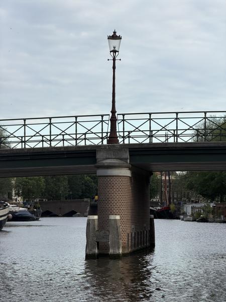This photo captures a serene view of a canal in Oudeschans, Uilenburg, Amsterdam, featuring a prominent lamppost on a brick pillar supporting a bridge